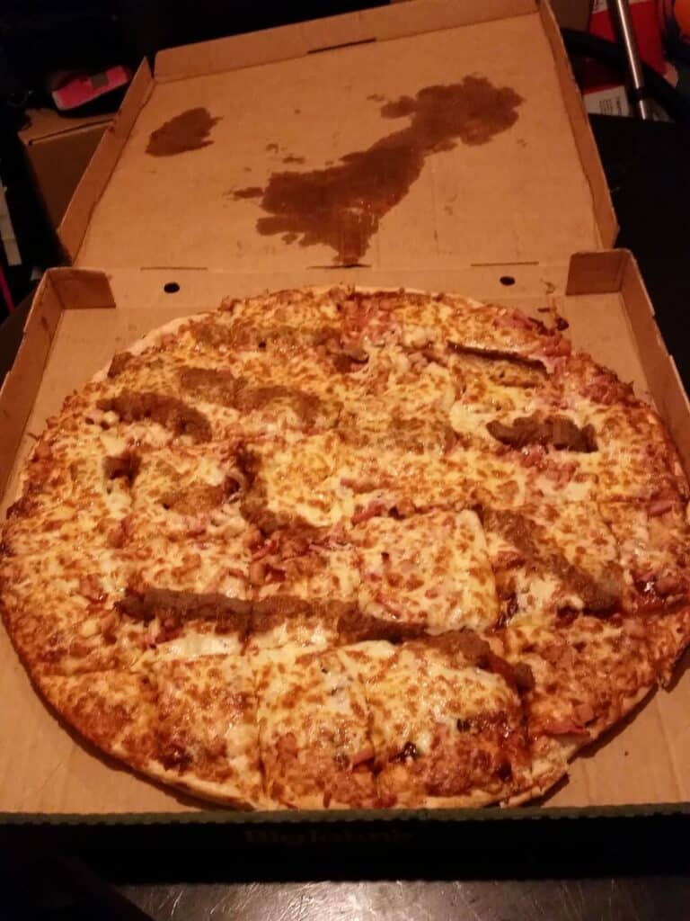 An open pizza box with a large pizza inside. The pizza is topped with cheese and several uneven strips of sausage. There is a large grease stain on the inside of the box lid, and the pizza appears slightly unevenly cut. The background is dark.