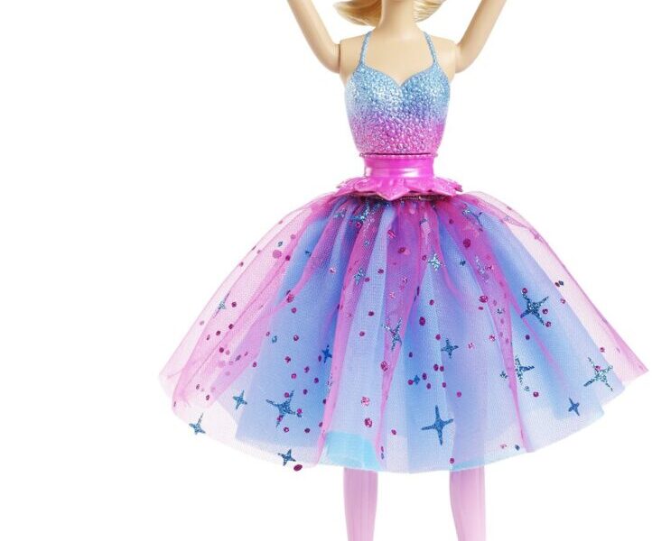 A Barbie doll dressed as a ballerina wears a blue and purple sparkly tutu, pink ballet shoes, and a pink tiara. Her arms are raised overhead in a ballet pose. A pink accessory tool lies on the ground nearby.