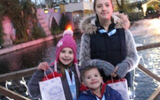 Three children stand on a wooden bridge decorated with fairy lights, holding festive gift bags. They wear winter jackets and hats. Behind them is a pond, trees, and Christmas lights, creating a cheerful outdoor, evening scene.