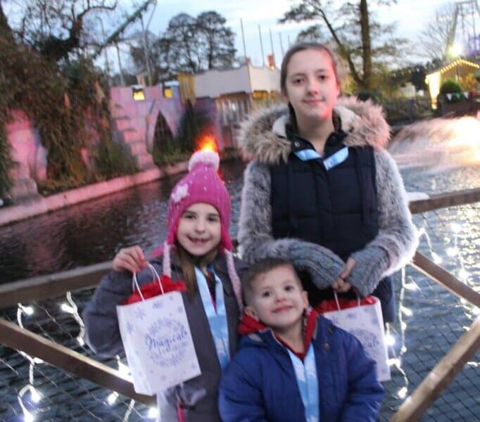 Three children stand on a wooden bridge decorated with fairy lights, holding festive gift bags. They wear winter jackets and hats. Behind them is a pond, trees, and Christmas lights, creating a cheerful outdoor, evening scene.