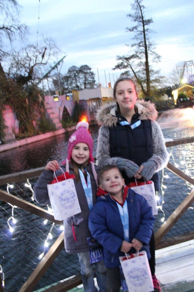 Three children stand on a wooden bridge decorated with fairy lights, each holding a festive gift bag. The eldest wears a grey coat with a fur hood, the middle child has a pink hat with a red pom-pom, and the youngest has a blue coat. A pond and trees are in the background.
