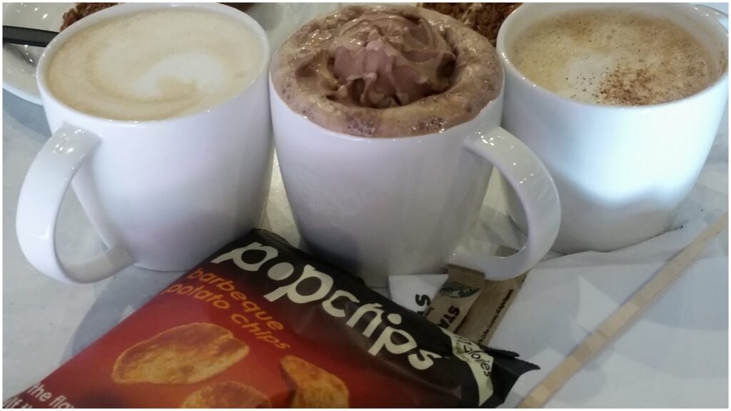 Three white mugs sit side by side: one with a latte, one with hot chocolate topped with whipped cream, and one with cappuccino. In front is a bag of Popchips barbecue crisps on a table, along with serviettes and sugar sachets.