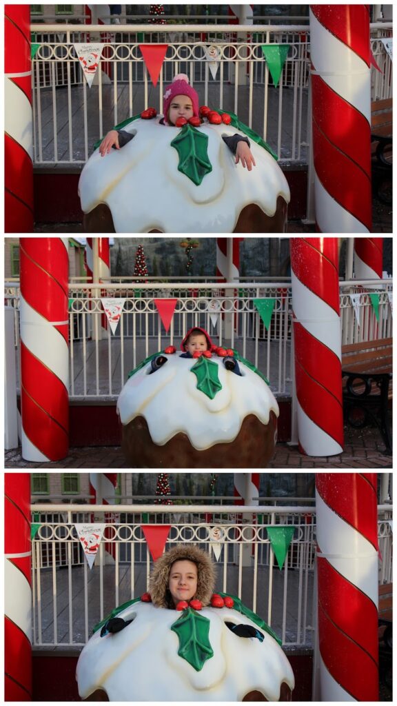 Three people pose separately behind a large, festive Christmas pudding display with white icing and holly leaves. Each person’s head sticks out of the pudding. Red-and-white striped poles and festive bunting decorate the background.