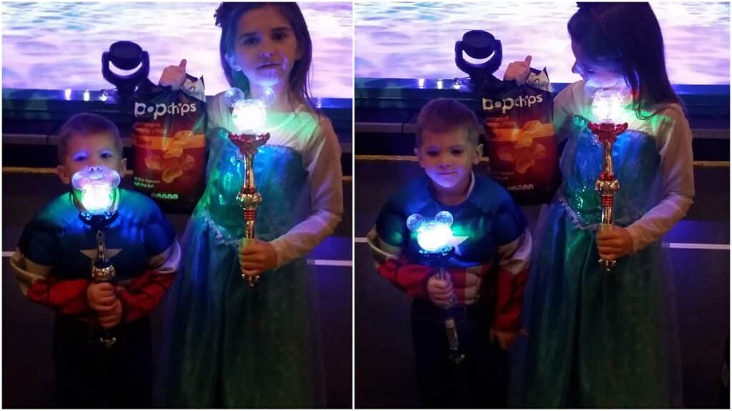Two children in fancy dress stand indoors holding glowing wands. The girl wears a blue princess dress and holds a bag of Popchips; the boy wears a superhero outfit. A bright, colourful light shines from their wands. The background is dim with a purple-lit area.