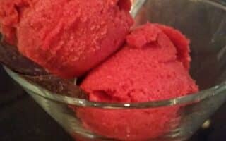 A glass bowl holds two scoops of bright red sorbet, served with a metal spoon. The surface beneath the bowl is dark, and there are chairs and tables blurred in the background, suggesting the setting is a café or restaurant.
