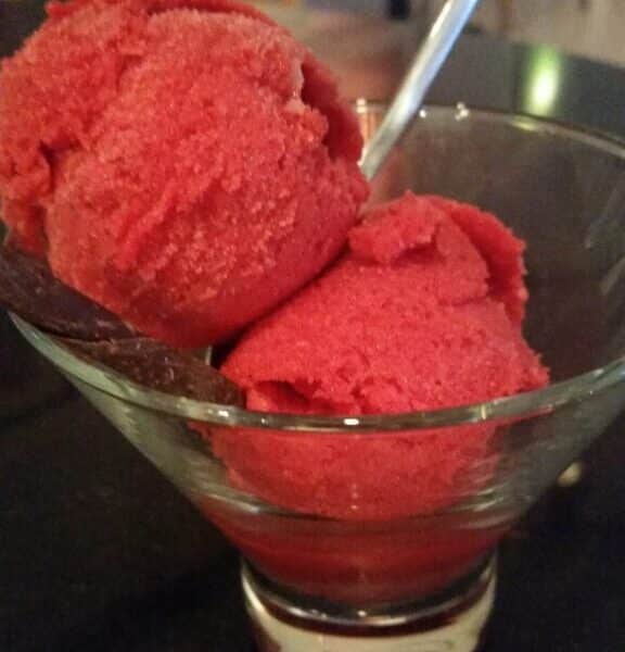 A glass bowl holds two scoops of bright red sorbet, served with a metal spoon. The surface beneath the bowl is dark, and there are chairs and tables blurred in the background, suggesting the setting is a café or restaurant.