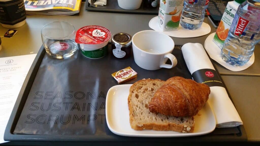 A breakfast tray with a croissant, two slices of multigrain bread, a pat of Président butter, a cup, glass, serviette with cutlery, a small jam jar, yoghurt, and juice; water and magazines are visible in the background.