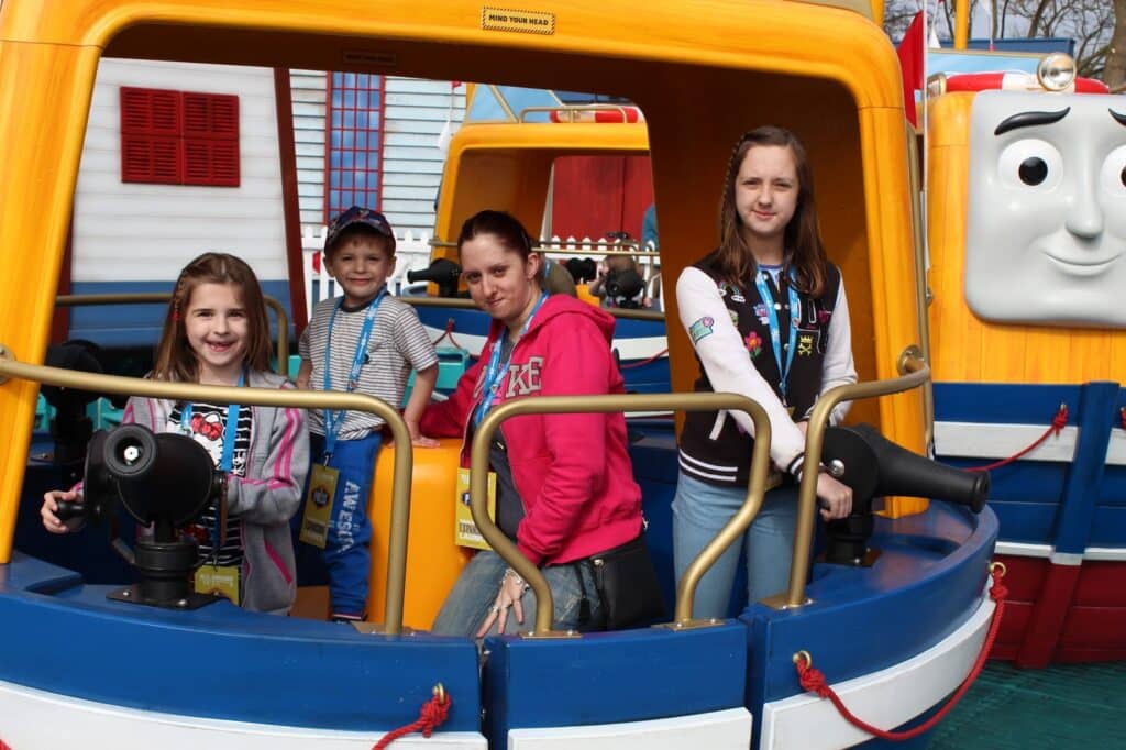 Four people—three children and one adult—pose on a blue and yellow cartoon-style boat ride. The boat features playful eyes and a face. All are smiling or looking at the camera. The background includes more colourful boats and amusement park scenery.