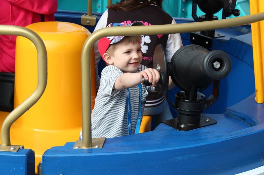 A young boy wearing a red cap and striped shirt smiles as he turns a black ship’s wheel on a blue and yellow playground structure, with other people visible in the background.
