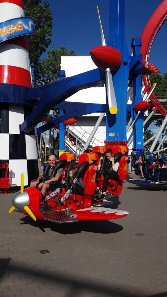 Three people, including an adult and two children, sit in a red, aeroplane-shaped amusement park ride. The ride has yellow wings and a propeller. In the background are colourful structures, including a striped tower and a looping rollercoaster.
