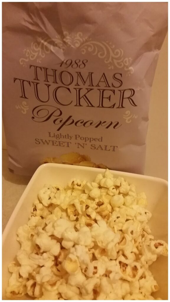 A bowl filled with popcorn sits in front of a light purple bag labelled “1988 Thomas Tucker Popcorn, Lightly Popped, Sweet ’n’ Salt.” The popcorn appears white and fluffy with some caramelised spots.