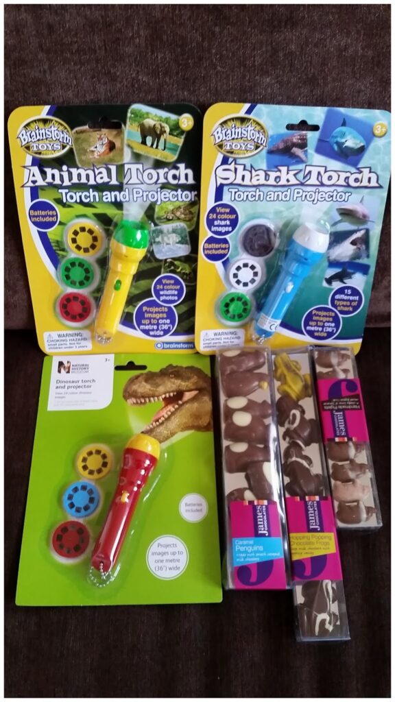 Three colourful toy torches in packaging—green (Animal Torch), blue (Shark Torch), and red (dinosaur theme)—each with picture reels. Beside them are three chocolate bars with animal patterns, all placed on a brown surface.