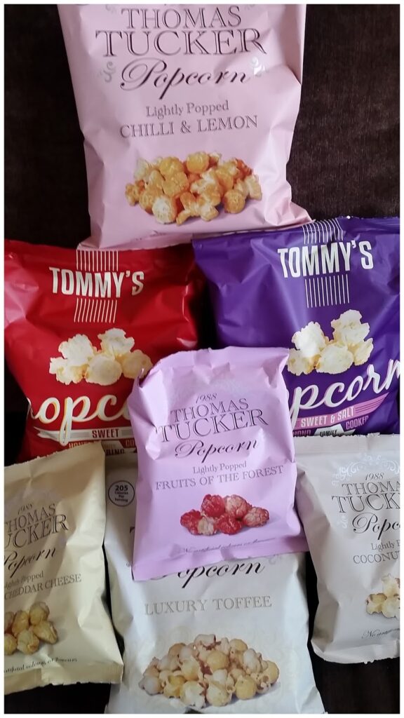 Seven packets of Thomas Tucker popcorn are arranged in a group. Flavours include: Lightly Popped Chilli & Lemon (pink), Sweet & Salty (red), Fruits of the Forest (purple), Cheddar Cheese (gold), Luxury Toffee (white), and Sweet & Crunchy (white).