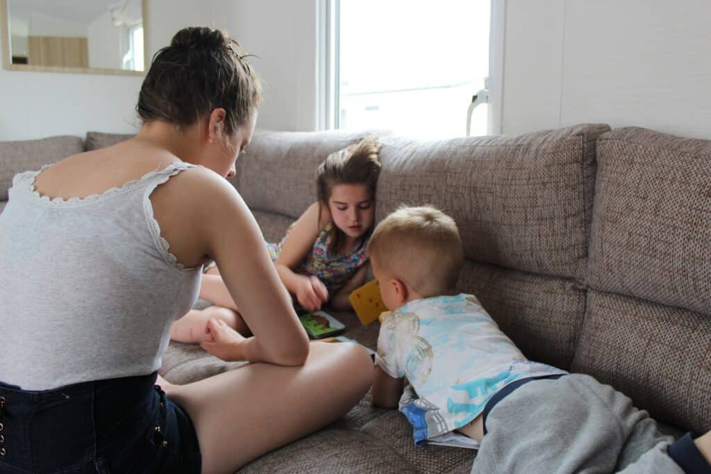 A woman and two children sit together on a beige sofa. The children, a girl and a boy, are focused on a book in front of them, while the woman, with her back to the camera, watches and engages with them in a bright, cosy room.