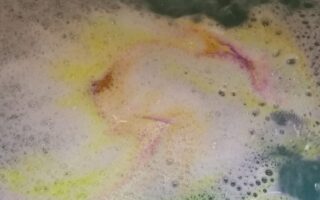 Colourful bathwater with bubbles in a tub. Swirls of yellow, pink, and orange mix in the water, creating a marbled effect, while a green area is visible in the top right. The scene suggests a dissolving bath bomb under running water.