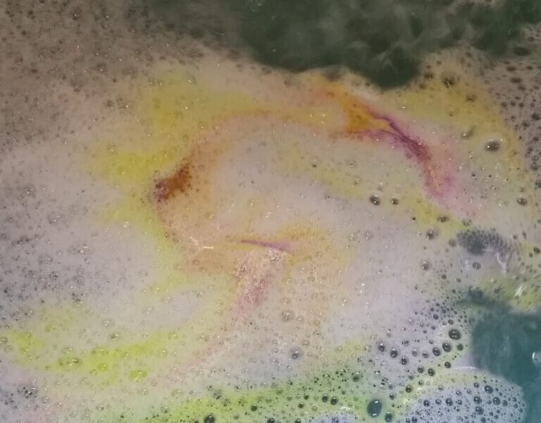 Colourful bathwater with bubbles in a tub. Swirls of yellow, pink, and orange mix in the water, creating a marbled effect, while a green area is visible in the top right. The scene suggests a dissolving bath bomb under running water.