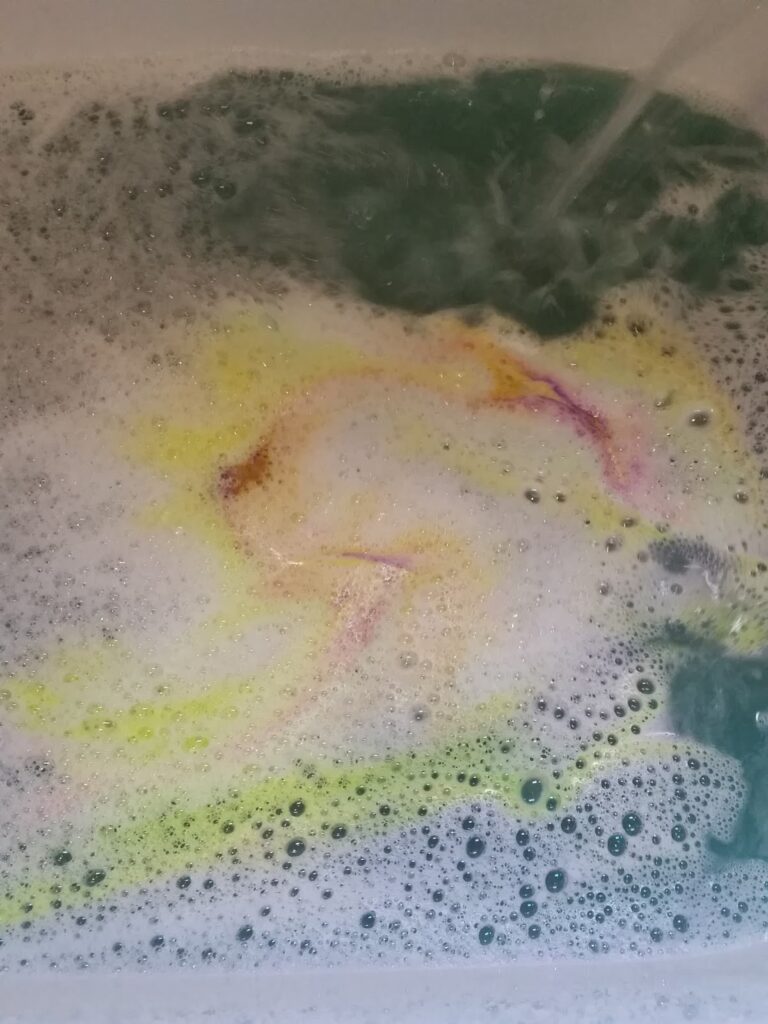 Water in a bath with white foam and swirling colours, including yellow, orange, pink, and green, spreading out as a bath bomb dissolves, creating a marbled effect on the water’s surface.