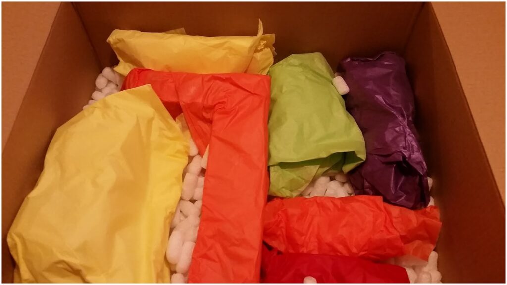 A cardboard box filled with crumpled yellow, orange, green, and purple tissue-wrapped items, surrounded by white packing peanuts. The colourful packages are assorted shapes and sizes, nestled securely within the box.