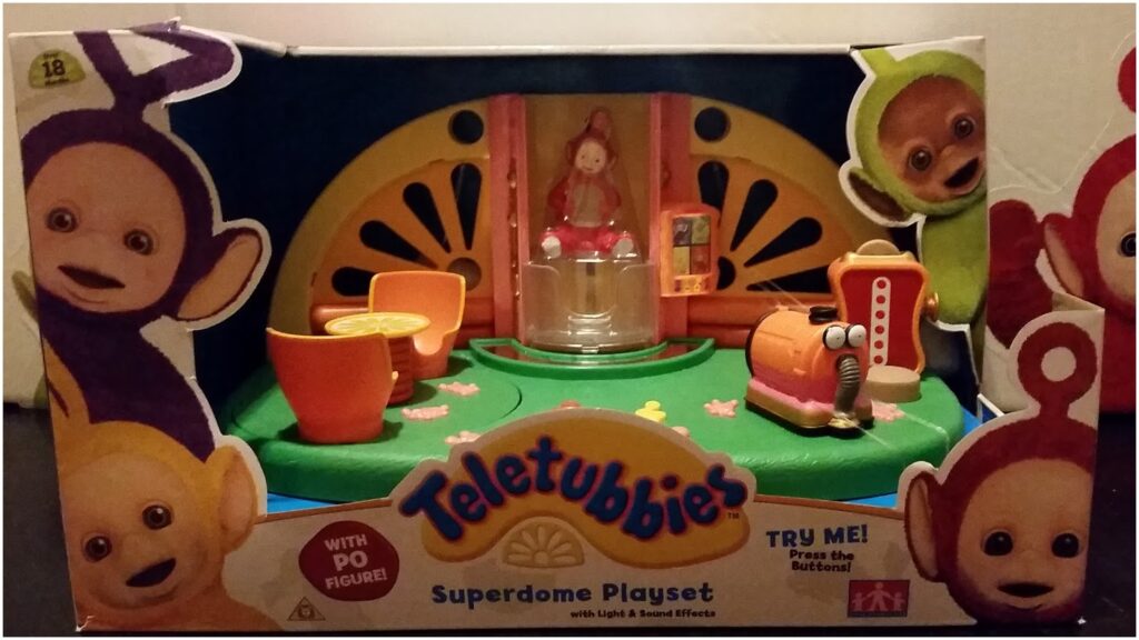 A Teletubbies Superdome Playset toy in its box features a round green base with orange tables, chairs, a vacuum cleaner, and a spinning Po figure in the centre. The box is decorated with pictures of all four Teletubbies and text promoting light and sound effects.