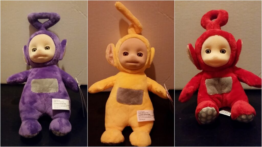 Three plush Teletubbies dolls are shown side by side: Tinky Winky (purple), Laa-Laa (yellow), and Po (red). Each has a sewn-on grey square on their tummy, unique head shapes, round ears, and pale faces with wide, round eyes.
