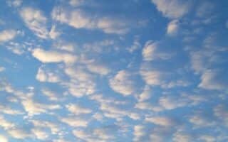 A sky filled with scattered, fluffy white clouds during daylight, creating a textured pattern. The light blue sky shows hints of sunlight, and silhouettes of rooftops, trees, and telegraph poles are visible along the bottom edge.