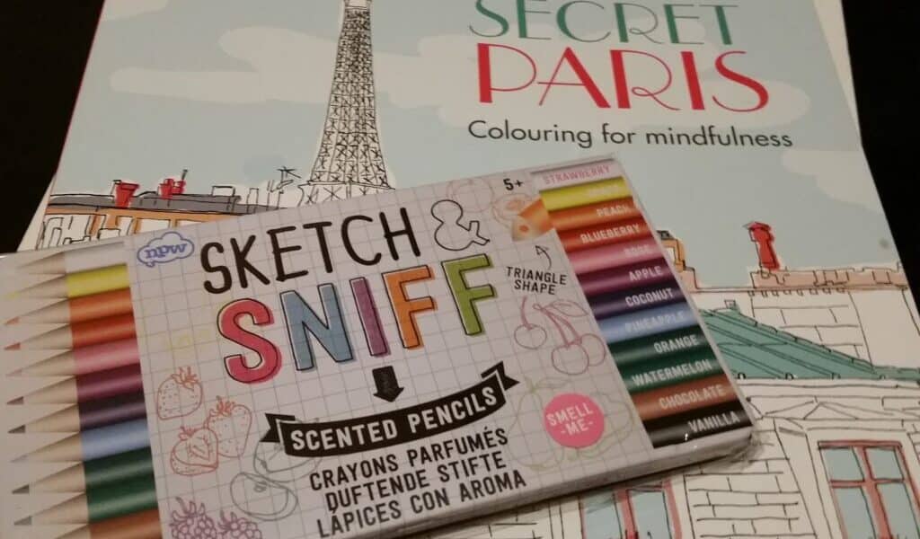 A box of colourful scented pencils labelled “Sketch & Sniff” lies on top of a colouring book titled “Secret Paris Colouring for mindfulness,” featuring an illustrated Paris scene with the Eiffel Tower and rooftops in pastel colours.