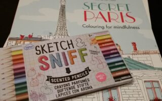 A box of colourful scented pencils labelled “Sketch & Sniff” lies on top of a colouring book titled “Secret Paris Colouring for mindfulness,” featuring an illustrated Paris scene with the Eiffel Tower and rooftops in pastel colours.