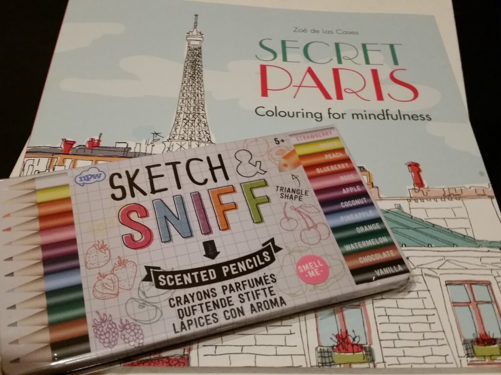 A pack of colourful scented pencils labelled "Sketch & Sniff" rests on a colouring book titled "Secret Paris" featuring an Eiffel Tower illustration and pastel rooftop scene. The pencils are arranged neatly, showing various scent names.