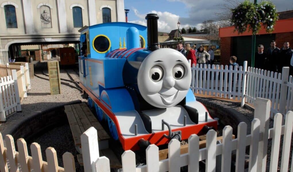 A life-sized model of Thomas the Tank Engine sits on display on a circular track, surrounded by a white picket fence. People stand nearby, and buildings, including one resembling a railway station, are visible in the background under a partly cloudy sky.