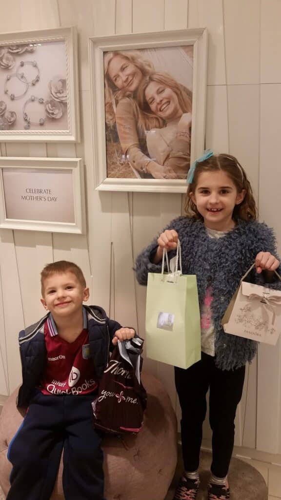 Two smiling children stand indoors holding shopping bags. Behind them is a wall with framed photos of two women and a child, plus a sign reading “Celebrate Mother’s Day.” The girl wears a blue hair bow and furry jacket; the boy wears a sports jacket and maroon shirt.