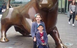 A young girl and boy smile while posing in front of a large bronze bull statue under a glass canopy. The area is paved with bricks, and a few people are visible in the background near shops and signs. The children appear happy and are close together.