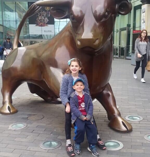 A young girl and boy smile while posing in front of a large bronze bull statue under a glass canopy. The area is paved with bricks, and a few people are visible in the background near shops and signs. The children appear happy and are close together.