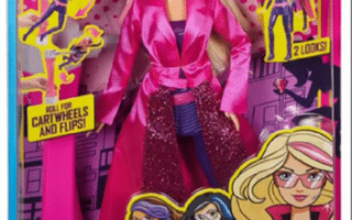 A boxed Barbie doll from the “Barbie Spy Squad” range wears a pink trench coat and sunglasses. The packaging is bright with cartoon spy graphics, featuring phrases like “Roll for cartwheels and flips!” and “2 looks.” For ages 3 and up; a warning is shown.