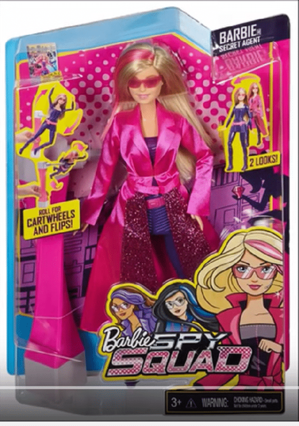 A boxed Barbie doll from the “Barbie Spy Squad” range wears a pink trench coat and sunglasses. The packaging is bright with cartoon spy graphics, featuring phrases like “Roll for cartwheels and flips!” and “2 looks.” For ages 3 and up; a warning is shown.