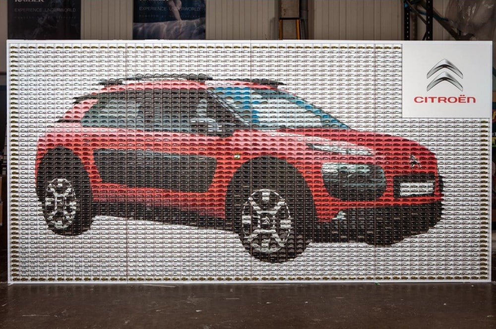 A mural made from hundreds of small tins arranged to depict a red Citroën car in profile. The Citroën logo and name appear on a white panel in the top right corner. The background is indoors with a warehouse-like setting.