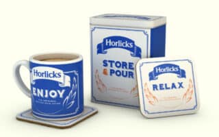 A branded Horlicks set with a blue and white design: a mug filled with a creamy drink on a matching coaster labelled "ENJOY," a square storage tin reading "STORE & POUR," and another coaster displaying "RELAX," all featuring laurel graphics.