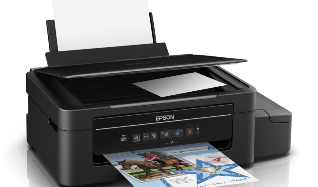 A black Epson all-in-one printer with a raised scanner lid, loaded with paper at the back. A colour document featuring images and text is being printed from the front tray. An external ink tank is visible on the right side of the printer.