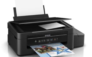 A black Epson all-in-one printer with a raised scanner lid, loaded with paper at the back. A colour document featuring images and text is being printed from the front tray. An external ink tank is visible on the right side of the printer.
