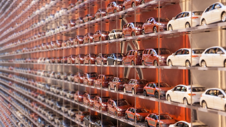 Rows of small model cars, mostly red and white, are neatly arranged on shelves that extend into the distance, creating a repeating, grid-like pattern. The image is taken from an angle, emphasising the depth and repetition of the miniature cars.