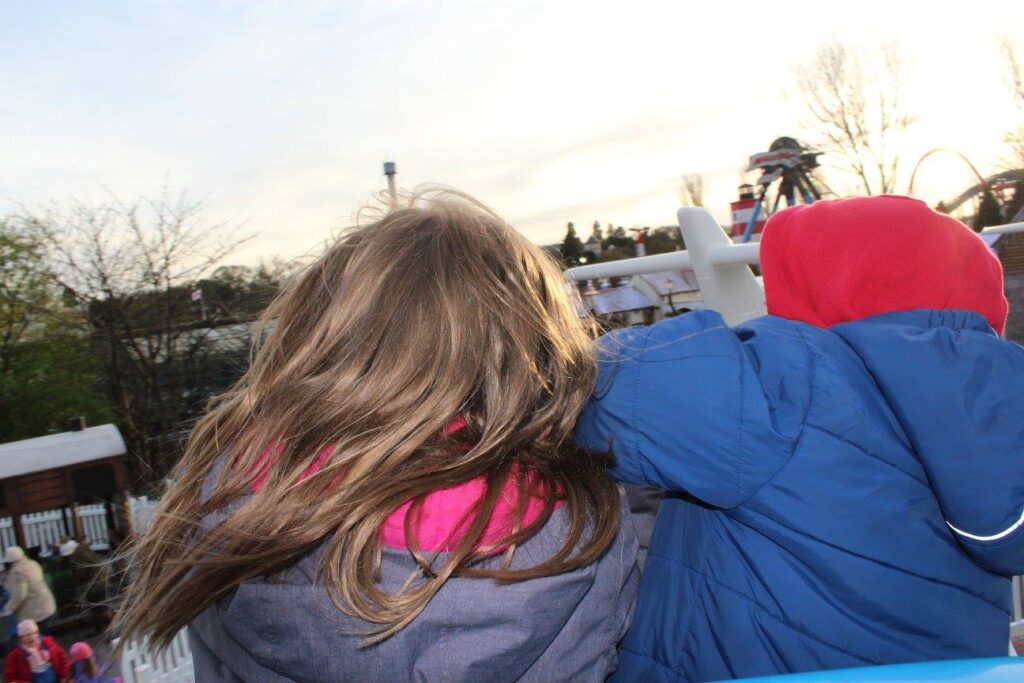 Two children, seen from behind, wear hooded jackets—one grey with brown hair visible, and one blue with a red hood. They lean over a white railing, looking towards a park or fairground with trees, people, and rides in the background at dusk.