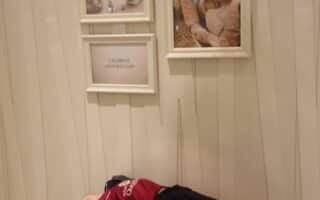 A young boy in casual clothes reclines on a round, tufted beige ottoman in a shop. Behind him are framed photos—two women smiling, jewellery pieces—and a sign that reads “CELEBRATE MOTHER’S DAY” on a cream-coloured patterned wall.