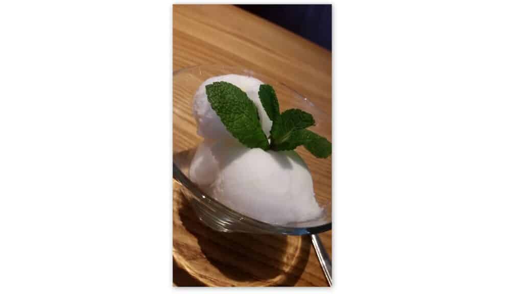 Two scoops of white sorbet are served in a small glass bowl, garnished with a fresh mint sprig. The bowl rests on a wooden surface, and the handle of a spoon is visible beside it. The lighting highlights the smooth texture of the sorbet.