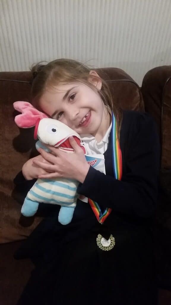 A young girl with light brown hair, wearing a navy cardigan and rainbow-coloured medal ribbon, smiles whilst hugging a colourful plush toy with pink ears, striped body, and one large eye, sitting on a brown sofa with a light background.