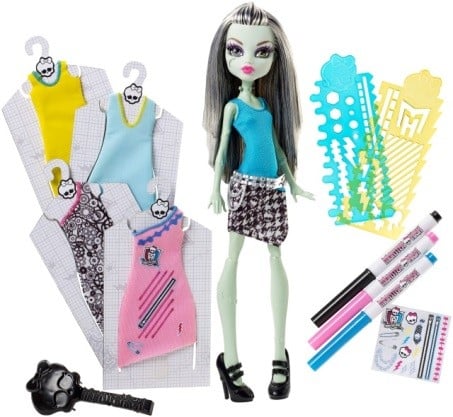 A green-skinned fashion doll with long black and white hair, wearing a blue top and patterned skirt, stands beside three extra outfits on hangers, three plastic stencils, two markers, a black hairbrush, and two small notepads with colourful designs.