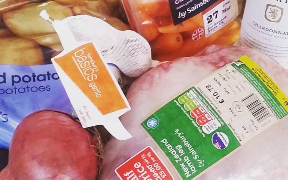 A close-up of groceries including a raw chicken with a green label, a bag of British baby Chantenay carrots, a netted bag of garlic, red potatoes, a red onion, and a bottle of McGuigan Estate Chardonnay wine.