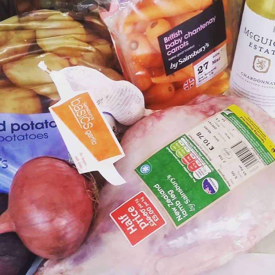 A close-up of groceries including a raw chicken with a green label, a bag of British baby Chantenay carrots, a netted bag of garlic, red potatoes, a red onion, and a bottle of McGuigan Estate Chardonnay wine.