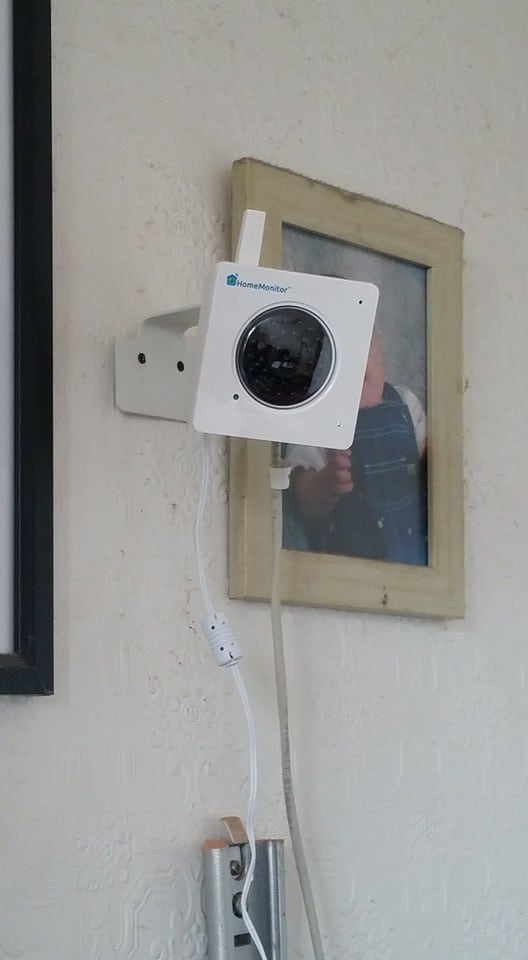 A white security camera is mounted on a wall, slightly tilted downwards, with cables attached. Behind the camera, a framed photo of a young child is partially obscured. The camera brand reads “HomeMonitor.” The wall has a subtle textured pattern.