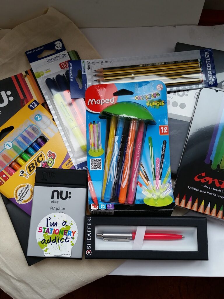 An assortment of stationery items including coloured pens, pencils, markers, a red and silver pen, paintbrushes, watercolour pencils, a notepad with "I'm a STATIONERY addict" on the cover, and a pack of BIC coloured pens arranged on a light surface.