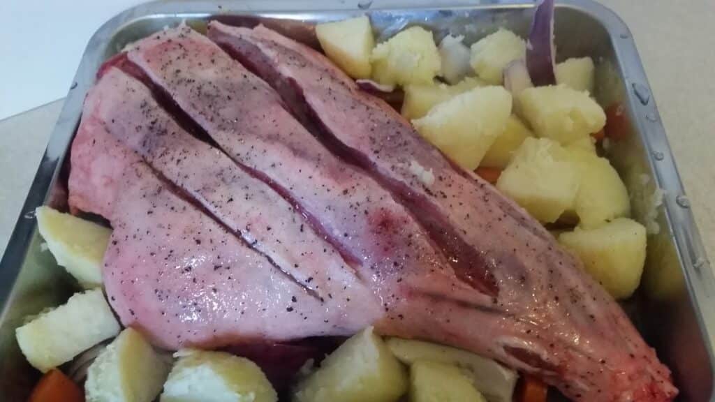 A raw seasoned joint of meat with three deep slashes on top rests on chopped potatoes and carrots in a metal roasting tin, ready to be cooked. The vegetables are cut into large pieces and surround the meat on all sides.