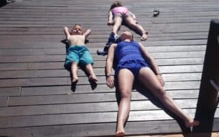 Three people, two children and one adult, lie on their backs on a sunlit wooden decking with their arms and legs relaxed, appearing to sunbathe. The scene is outdoors near tiled ground, with shadows and buildings in the background.